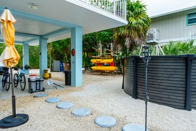 Tropical backyard scene with a light-blue covered patio, folded yellow umbrella, bicycles and fishing gear, stacked yellow kayaks on a rack, round above-ground pool and gravel with circular stepping stones.