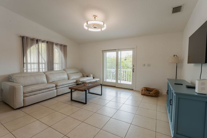 Bright living room with white leather sectional, wooden coffee table, sliding glass balcony doors, tiled floor, blue media console, floor lamp and small pet bed.