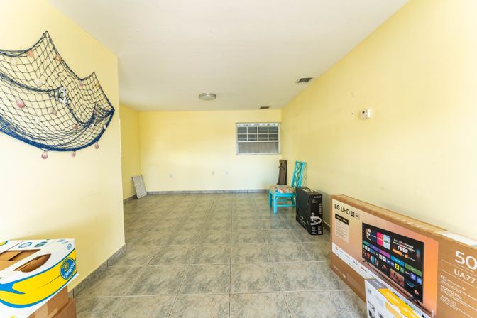 Empty beachy room with pale yellow walls and gray tile floor, decorative fishing net on left wall, small window, teal chairs stacked near boxes and a boxed flat-screen TV along the right wall.