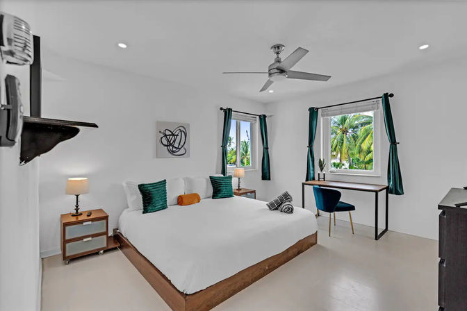 Bright modern tropical bedroom with a king-size bed, teal curtains and pillows, wooden nightstands and desk, ceiling fan, and windows framing lush palm-tree views — vacation rental vibe.