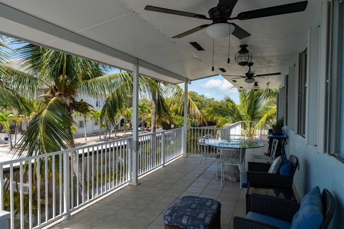 Shaded waterfront balcony with ceiling fans, wicker seating and glass dining table overlooking coconut palms and a calm canal — relaxing tropical outdoor patio scene.