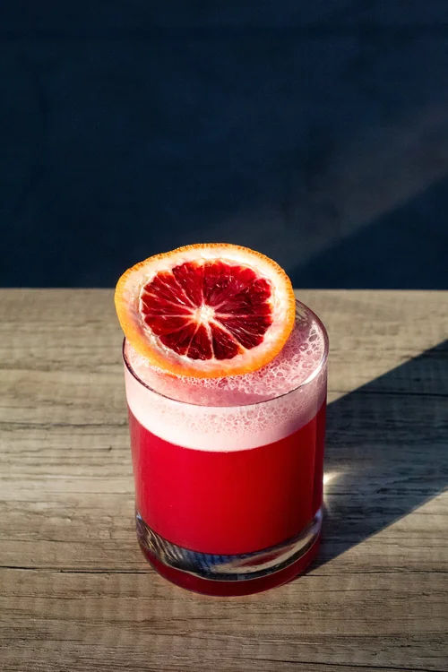 Vibrant blood orange cocktail in a lowball glass with a frothy pink head and a blood orange wheel garnish, sitting on a sunlit wooden table.