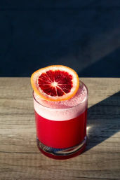Vibrant blood orange cocktail in a lowball glass with a frothy pink head and a blood orange wheel garnish, sitting on a sunlit wooden table.