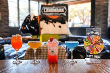 Vibrant orange, yellow, pink and blue cocktails lined up on a wooden table in a trendy bar, with a colorful spin-the-wheel shot menu and bull-themed decor blurred in the background.