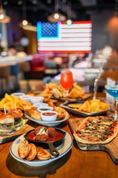 Colorful spread of American bar food on a wooden table — meatballs in a cast-iron skillet, flatbread pizza, sliders, chips with dips and bright cocktails, blurred neon American flag in the background