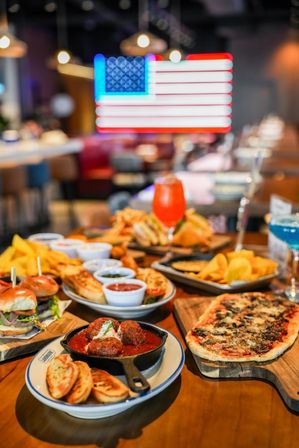 Colorful spread of American bar food on a wooden table — meatballs in a cast-iron skillet, flatbread pizza, sliders, chips with dips and bright cocktails, blurred neon American flag in the background