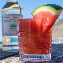 Sunlit watermelon cocktail in a textured rocks glass with a watermelon wedge garnish, set on a weathered wooden table with a clear liquor bottle and ocean cliffs in the blurred beach background.