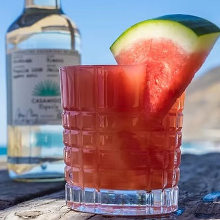Sunlit watermelon cocktail in a textured rocks glass with a watermelon wedge garnish, set on a weathered wooden table with a clear liquor bottle and ocean cliffs in the blurred beach background.