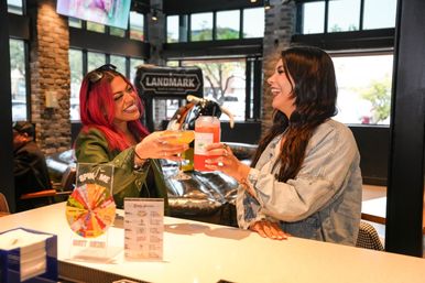 Two friends laughing and clinking bright cocktails at a modern downtown cocktail bar with stone accents, large windows, and a promotional spin-the-wheel.