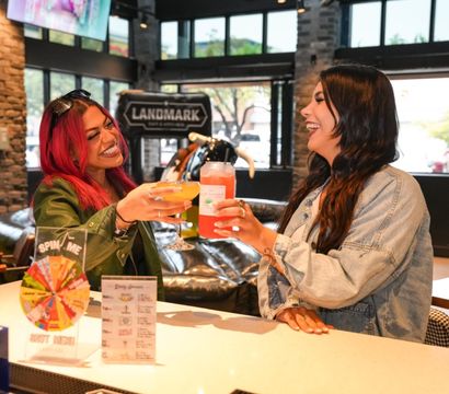 Two friends laughing and clinking bright cocktails at a modern downtown cocktail bar with stone accents, large windows, and a promotional spin-the-wheel.