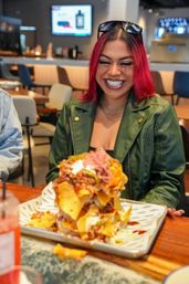 Beaming woman with bright pink hair and sunglasses on her head in a green jacket at a casual restaurant, excitedly eyeing a towering tray of loaded nachos with cheese, pickled onions and sour cream on a wooden table.