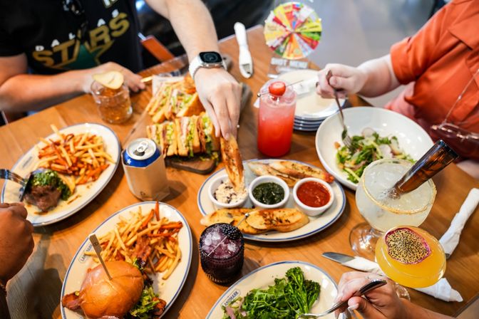 Overhead shot of a lively casual dining table with burgers and fries, stacked club sandwiches, salads, dipping sauces and colorful cocktails as hands reach in to share.