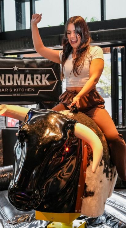 Young woman riding a mechanical bull indoors at a bar-style restaurant, one arm raised as she balances on a black-and-white faux bull inside an inflatable safety ring with brick walls and large windows.
