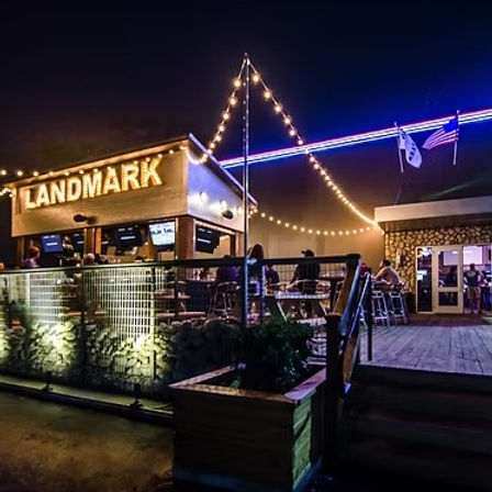 Outdoor bar and restaurant patio at night with warm string lights, neon roof trim, diners seated on a wooden deck and potted planters creating a cozy nightlife vibe.