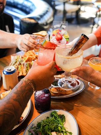 Diners clinking colorful cocktails over a wooden restaurant table at brunch — margarita with an upside-down beer bottle, pink watermelon-rimmed drink, blackberry cocktail, plus sandwiches, fries and a salad.