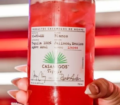 Hand with manicured nails holding a clear jar-style glass of bright red strawberry cocktail with ice, a strawberry garnish and white label, neon red, white and blue striped lights in the background.