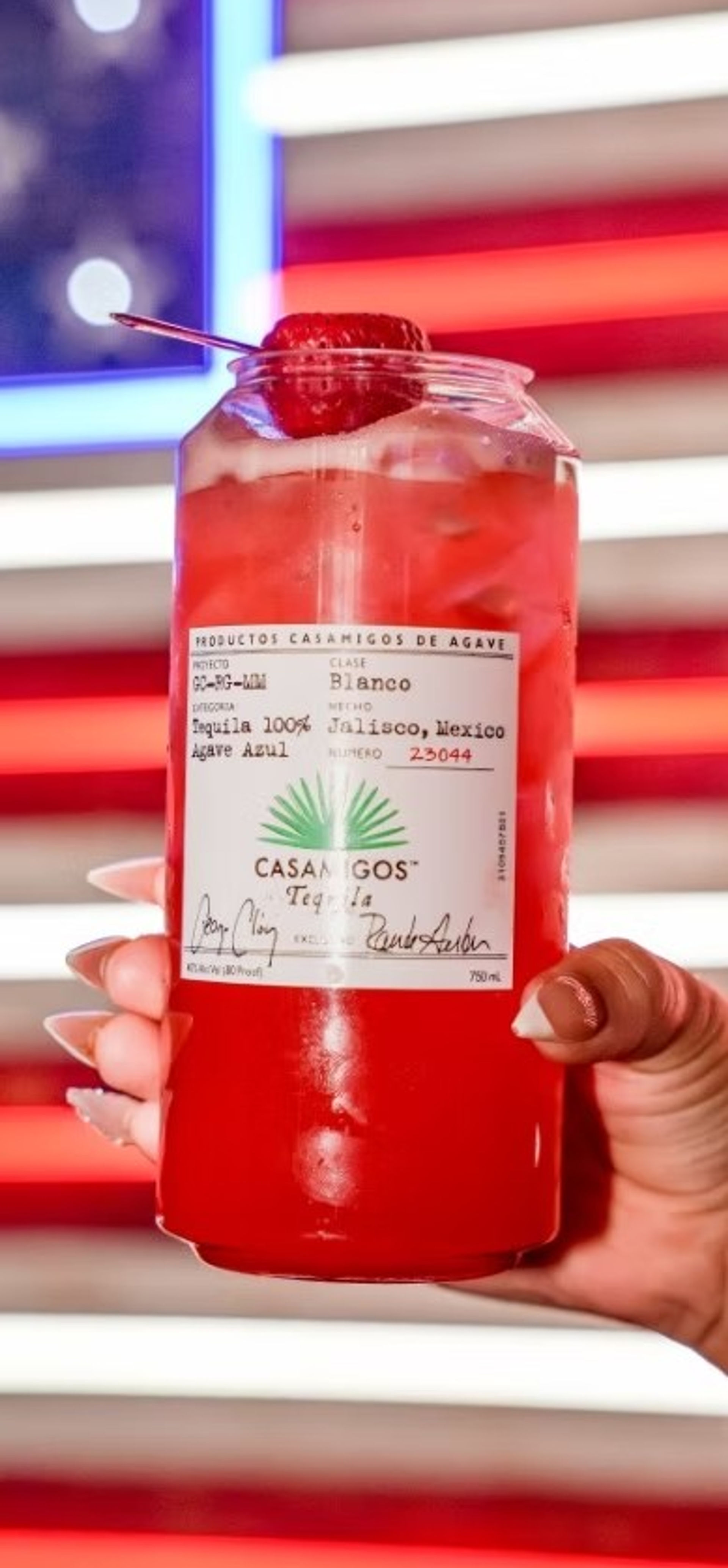 Hand with manicured nails holding a clear jar-style glass of bright red strawberry cocktail with ice, a strawberry garnish and white label, neon red, white and blue striped lights in the background.