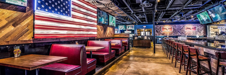 Wide-angle interior of an American sports bar with illuminated oversized US flag on the wall, red leather booths, a long wooden bar with high stools, multiple ceiling TVs, herringbone wood accents and polished concrete floor.