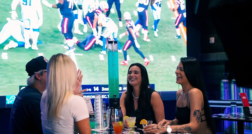 Group of friends at a lively sports bar watching American football on a giant screen, laughing around a table with a tall beer tower and colorful cocktails — game-day atmosphere.