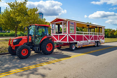 Bright red tractor towing a red-and-white open-air sightseeing wagon parked on a sunny downtown street beside a tree-lined park under a blue sky