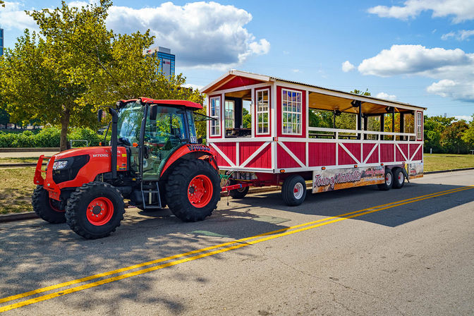 Bright red tractor towing a red-and-white open-air sightseeing wagon parked on a sunny downtown street beside a tree-lined park under a blue sky