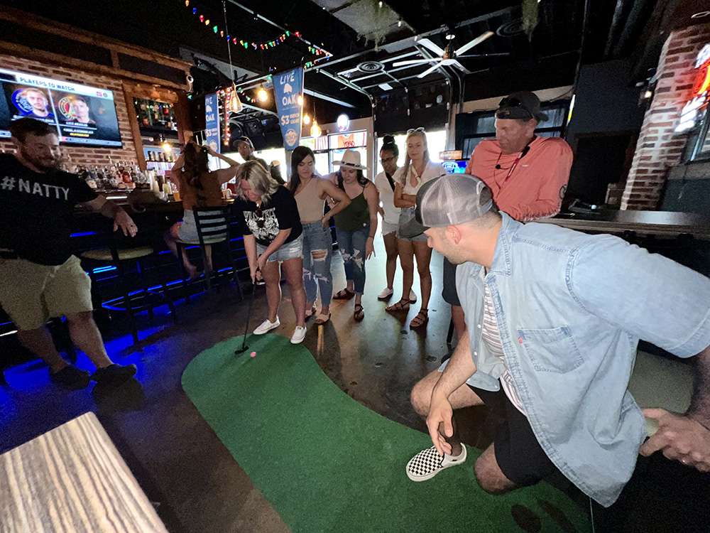 Group of friends playing on a small indoor putting green at a lively sports bar with neon lights, TVs, exposed brick, and a casual crowd gathered to watch.