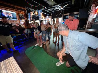 Group of friends playing on a small indoor putting green at a lively sports bar with neon lights, TVs, exposed brick, and a casual crowd gathered to watch.