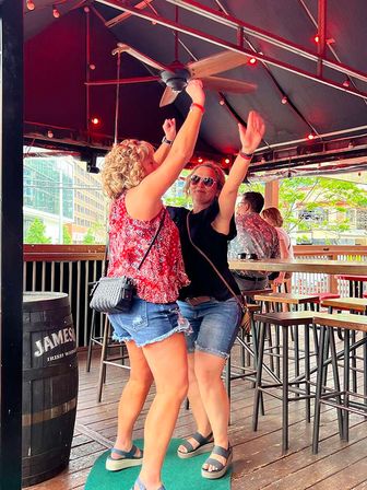 Two friends in shorts and sandals dancing under a ceiling fan on a covered urban rooftop patio bar with string lights, wooden deck, high-top tables and city buildings visible beyond.