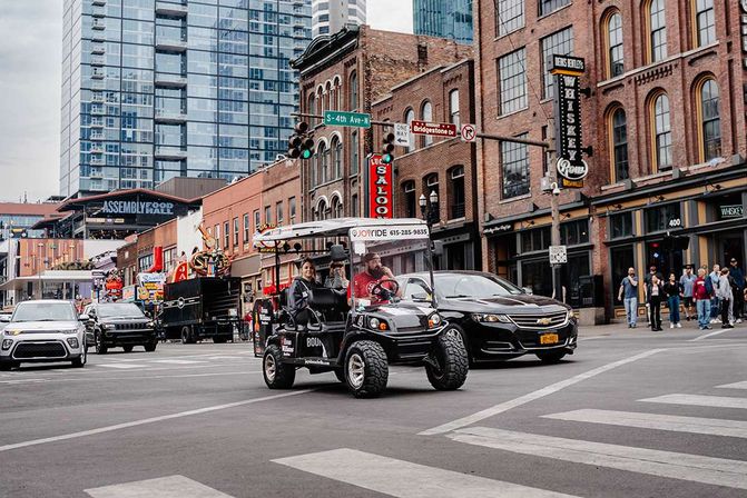 Small tour cart with passengers crossing a busy downtown intersection amid brick storefronts, neon signs, cars and pedestrians with glass high-rises in the background