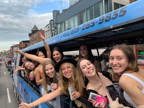 Smiling young women leaning out of a blue party trolley on a busy downtown city street, holding drinks and phones and waving during a daytime celebration
