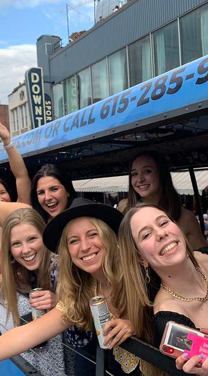 Smiling young women leaning out of a blue party trolley on a busy downtown city street, holding drinks and phones and waving during a daytime celebration
