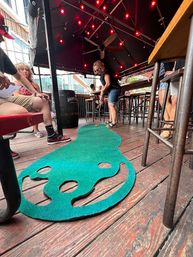 Outdoor bar patio on a wooden deck with red string lights and patrons playing on a green portable mini-golf putting mat.