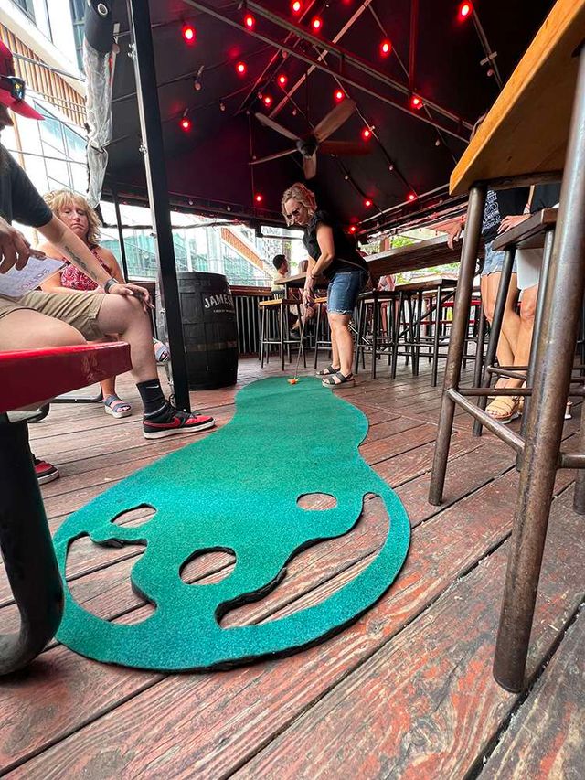 Outdoor bar patio on a wooden deck with red string lights and patrons playing on a green portable mini-golf putting mat.