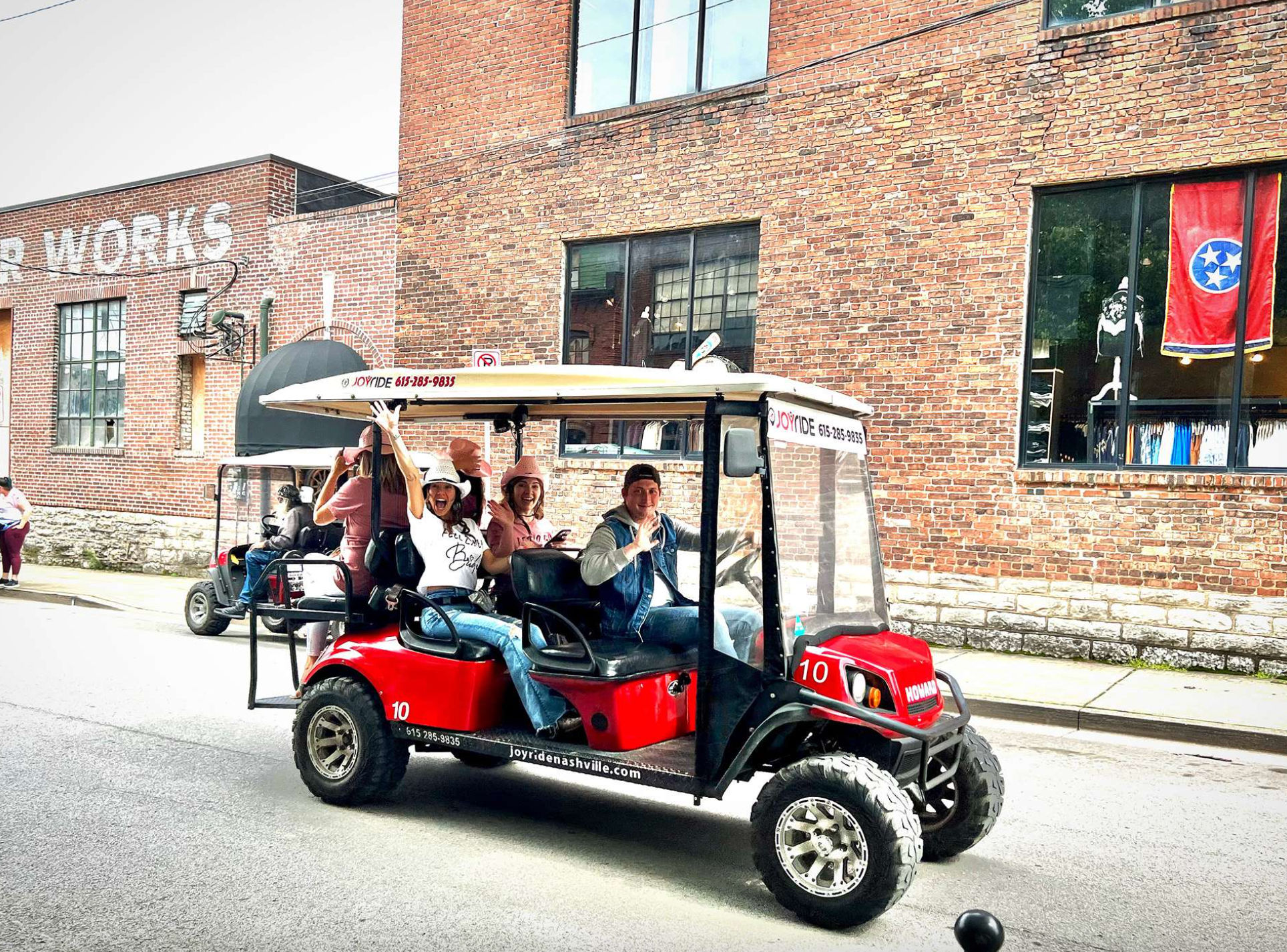 Smiling group waving from a red open-air tour cart rolling past brick storefronts and a Tennessee state flag in downtown Nashville.
