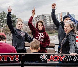 Smiling group of people in maroon college sweatshirts cheering and raising hands on a parade float with an American flag and urban skyline in the background.