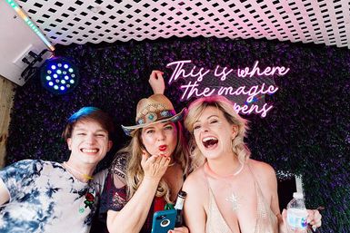 Three friends laughing in a colorful photo booth with a leafy backdrop and pink neon sign reading "This is where the magic happens" — woman in a cowboy hat blowing a kiss, a friend in a sparkly top holding a water bottle, and a smiling young man.