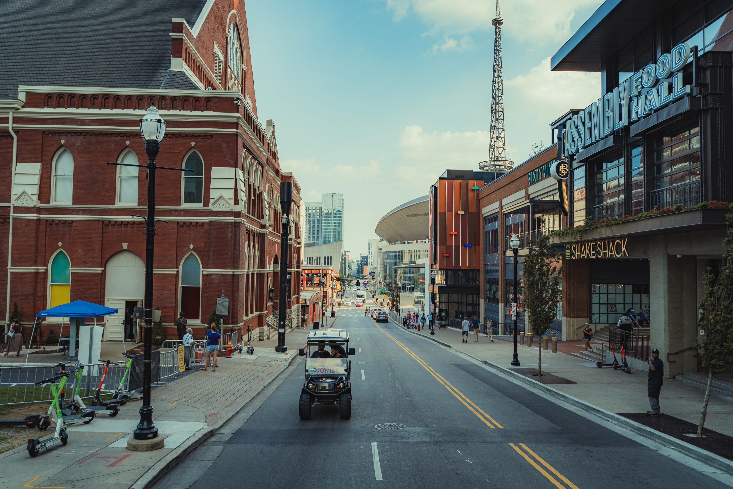 Downtown entertainment district: historic red-brick concert hall on the left, modern arena and mixed-use buildings on the right, scooters on the sidewalk, a utility cart driving down the street and pedestrians nearby.