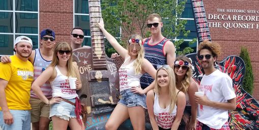 Group of young adults in patriotic summer outfits posing with oversized painted guitar sculptures in front of a brick music landmark at a sunny street festival.