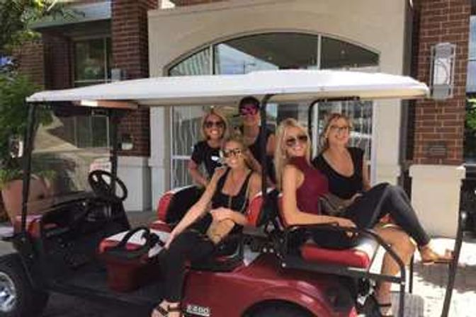 Five women in sunglasses smiling and posing on a red golf cart parked outside a brick building entrance on a sunny day.