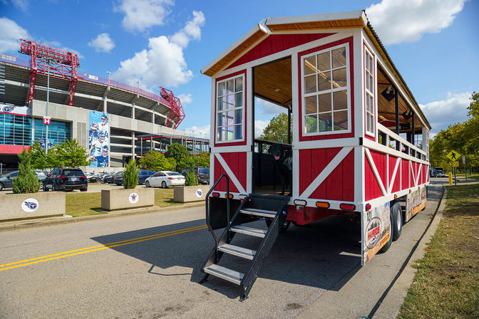Red-and-white open-air tailgate trolley with folding stairs parked on a sunny game-day street outside a large football stadium, with cars in the lot and a bright blue sky.