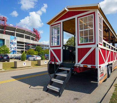 Red-and-white open-air tailgate trolley with folding stairs parked on a sunny game-day street outside a large football stadium, with cars in the lot and a bright blue sky.