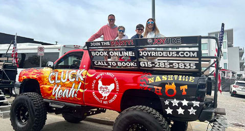 Lifted party pickup truck with colorful “Cluck Yeah” and Nashville hot-chicken graphics, four people riding in the bed on a city street.