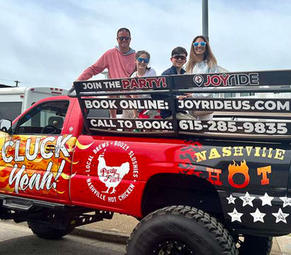 Lifted party pickup truck with colorful “Cluck Yeah” and Nashville hot-chicken graphics, four people riding in the bed on a city street.