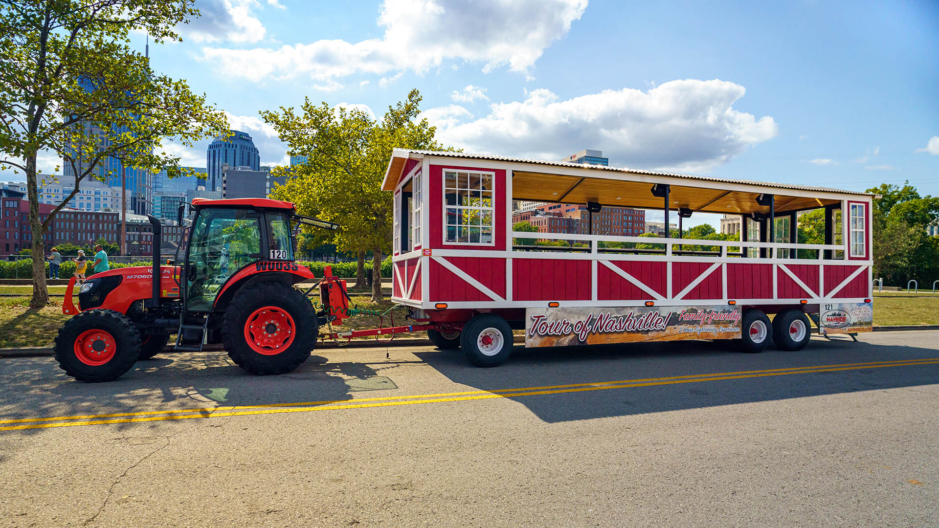 Red open-air trolley-style tour trailer reading 'Tour of Nashville' hitched to an orange tractor parked on a sunny city street with trees and the Nashville skyline in the background.