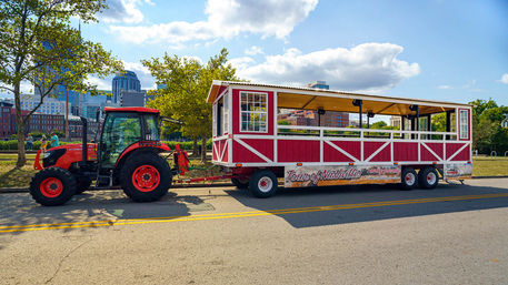 Red open-air trolley-style tour trailer reading 'Tour of Nashville' hitched to an orange tractor parked on a sunny city street with trees and the Nashville skyline in the background.