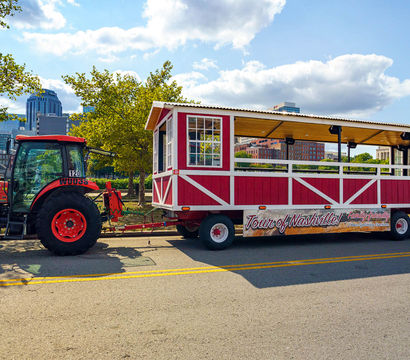 Red open-air trolley-style tour trailer reading 'Tour of Nashville' hitched to an orange tractor parked on a sunny city street with trees and the Nashville skyline in the background.