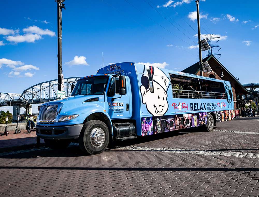 Bright blue open-air party bus with a smiling cartoon logo parked on a brick riverfront near a steel bridge under a sunny blue sky.