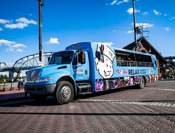 Bright blue open-air party bus with a smiling cartoon logo parked on a brick riverfront near a steel bridge under a sunny blue sky.