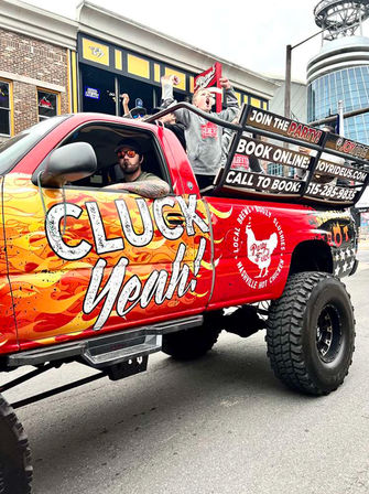 Red lifted party truck with flame 'Cluck Yeah' graphics and oversized tires, passengers cheering in the open bed while parked on a downtown street outside bars and restaurants.