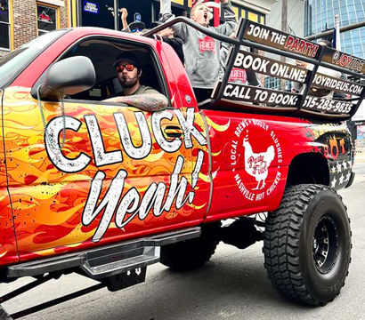 Red lifted party truck with flame 'Cluck Yeah' graphics and oversized tires, passengers cheering in the open bed while parked on a downtown street outside bars and restaurants.
